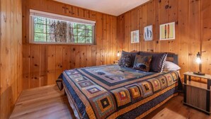 A wooden-paneled bedroom with a large bed, a window with a view of trees, and three framed pictures on the wall. - Triple C, Private Hot Tub, Dog-Friendly (Carnelian Bay)