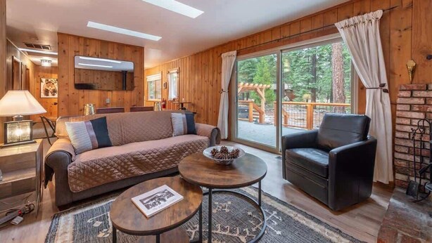 A cozy room with a wooden interior, a sofa, a chair, a table with a bowl, and a large window with a view of the outdoors. - Triple C, Private Hot Tub, Dog-Friendly (Carnelian Bay)