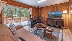A cozy room with a fireplace, a sofa, a coffee table, and a large window with a view of trees and a cabin. - Triple C, Private Hot Tub, Dog-Friendly (Carnelian Bay)