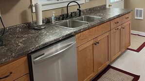 A kitchen with wooden cabinets, a granite countertop, a dishwasher, and a window with plaid curtains. - Millheim Mallard Place (Millheim)