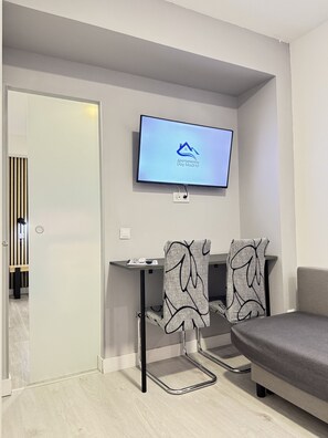Uma sala de estar moderna com uma TV de tela plana na parede exibindo 'Apartamentos Day Madrid'. Há um sofá e duas poltronas estampadas ao redor de uma mesa pequena com um controle remoto.