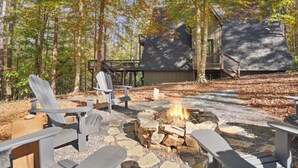 Eine Holzhütte mit einem Steinkamin, umgeben von Adirondack-Stühlen, inmitten eines bewaldeten Geländes mit einer Terrasse und einer Treppe, die zur Hütte führt.