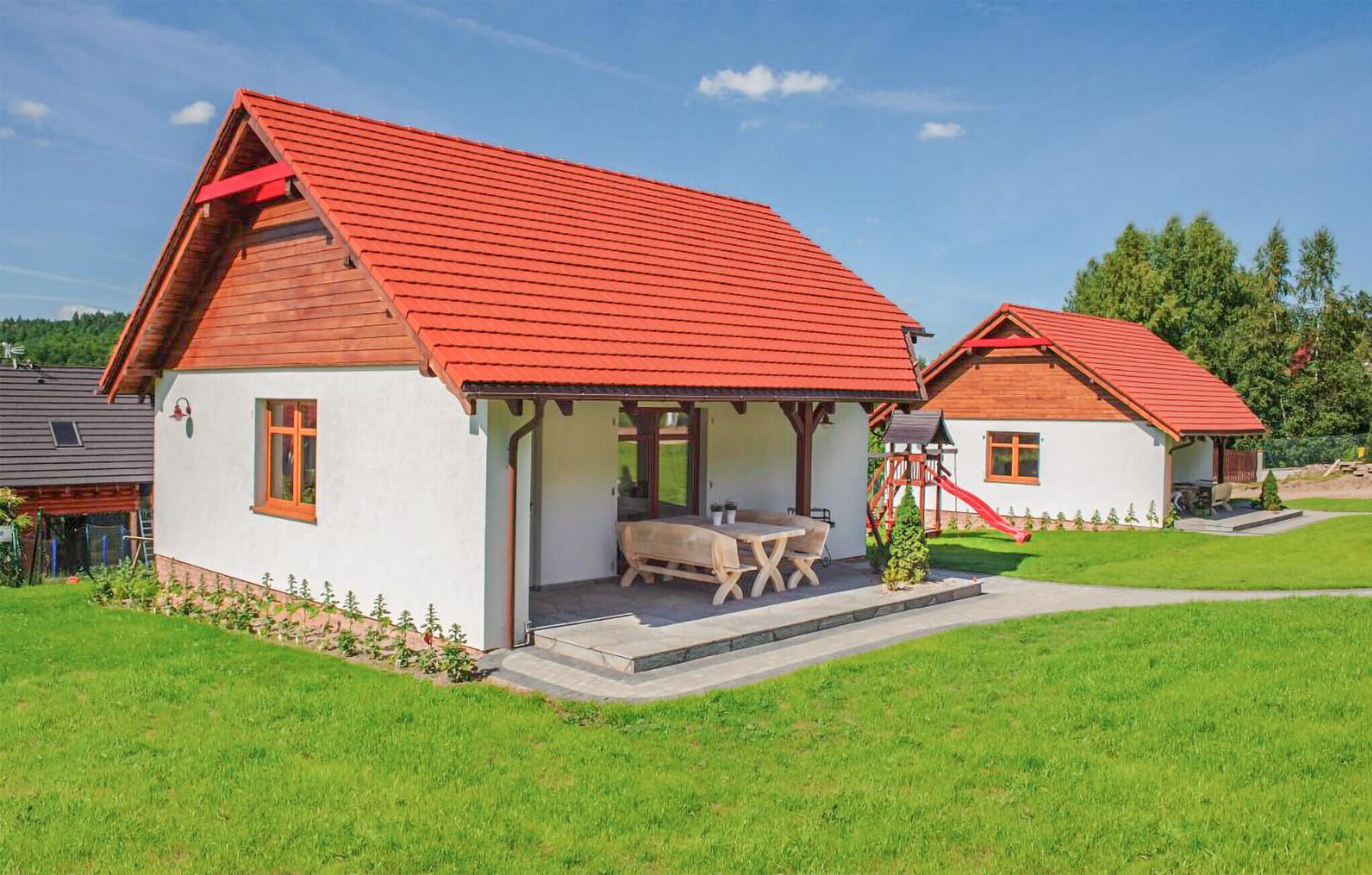 A two-story house with a red roof, a covered patio with a table and chairs, and a small play area with a slide.