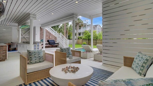 An outdoor patio area with wicker furniture, a round table, and a view of a backyard with a pool and stairs leading to a house. - Be a Sullivans Local! 1307 Cove Avenue by Avantstay Pool + Outdoor Living (Sullivan's Island)