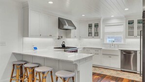 A modern kitchen with white cabinetry, a central island with stools, stainless steel appliances, and a window with a view. - Be a Sullivans Local! 1307 Cove Avenue by Avantstay Pool + Outdoor Living (Sullivan's Island)