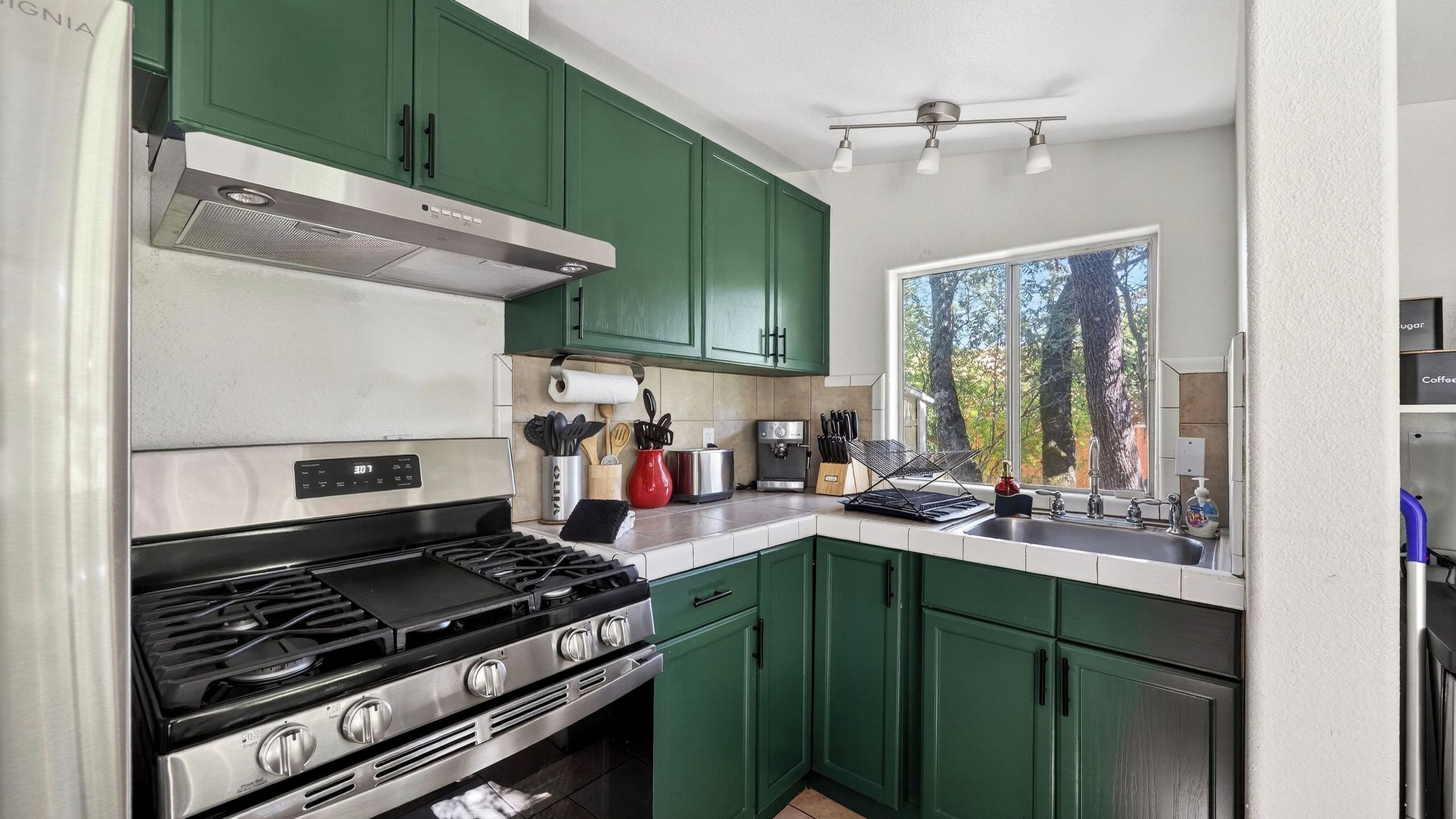 Une cuisine avec des meubles de couleur verte, une cuisinière, une machine à café et une fenêtre donnant sur des arbres.