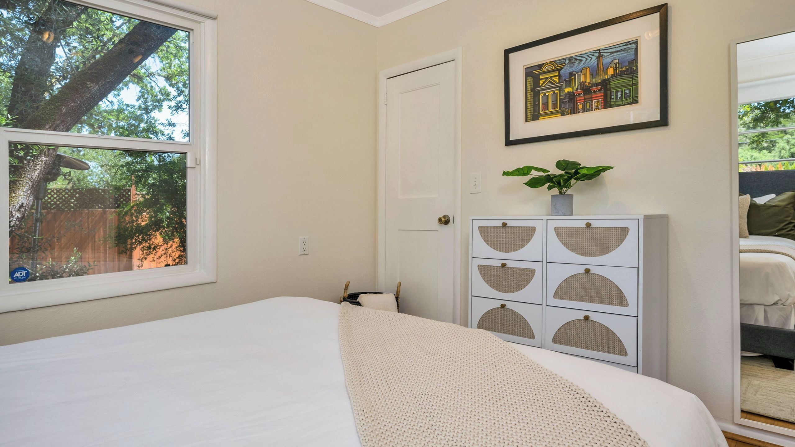 A bedroom with a bed, a door, a framed cityscape picture, a plant on a cabinet, and a window with a view of trees.