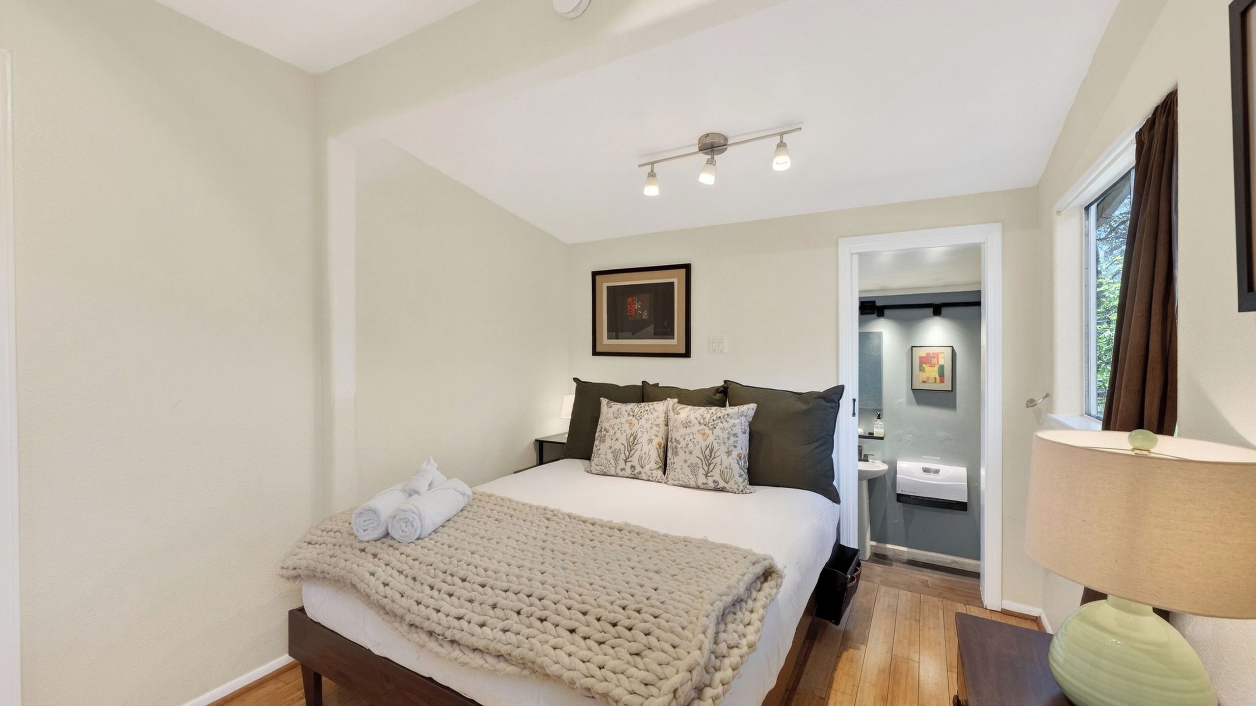 A bedroom with a bed, bedside tables, a window, and a bathroom visible through an open door.