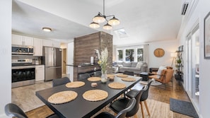 Una cocina moderna con electrodomésticos de acero inoxidable, un comedor con una mesa oscura y individuales tejidos, y una sala de estar con un sofá y un gran reloj de pared.