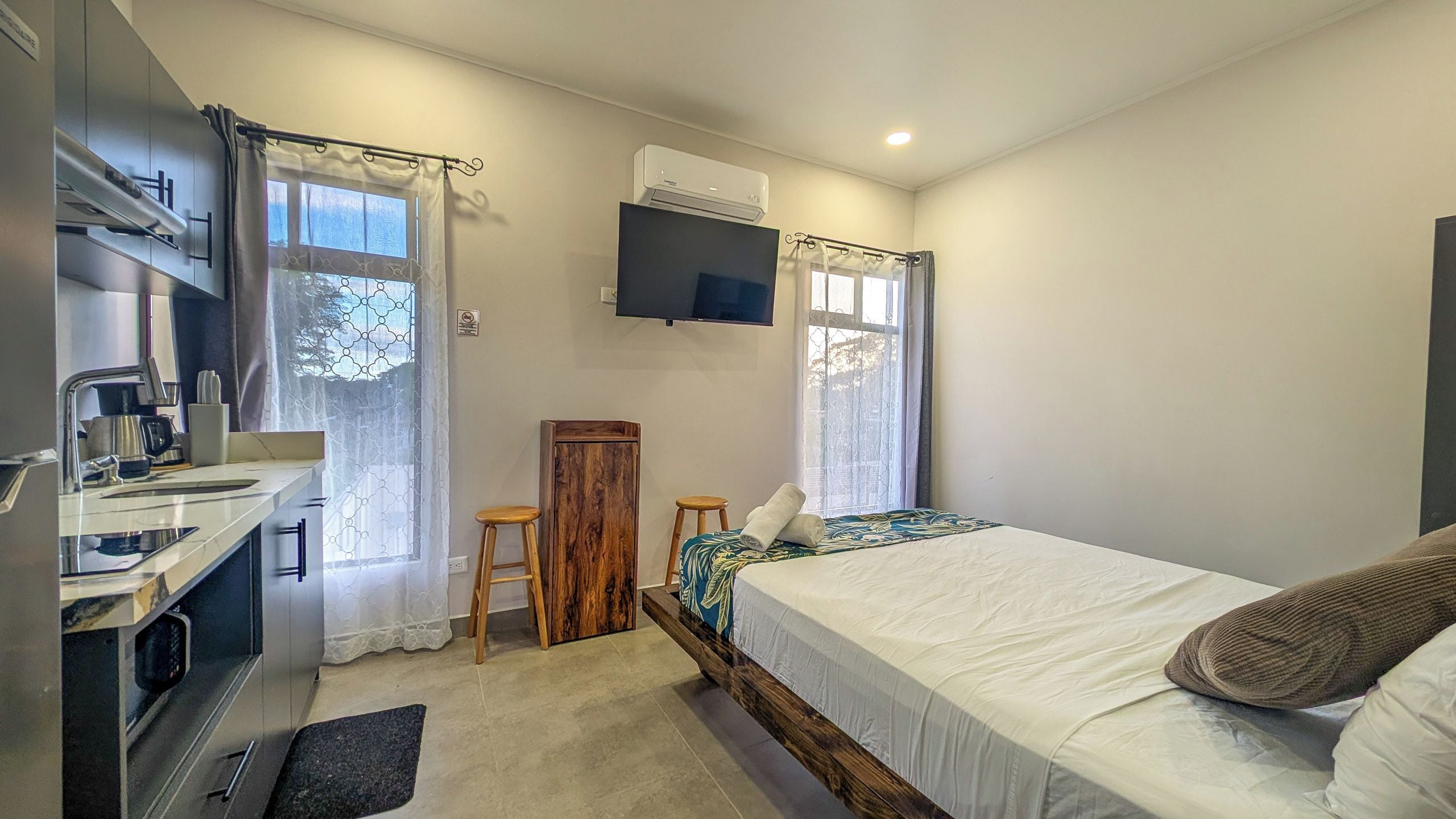 A compact hotel room with a bed, a desk with a chair, a small stool, a wooden cabinet, and a mounted TV.