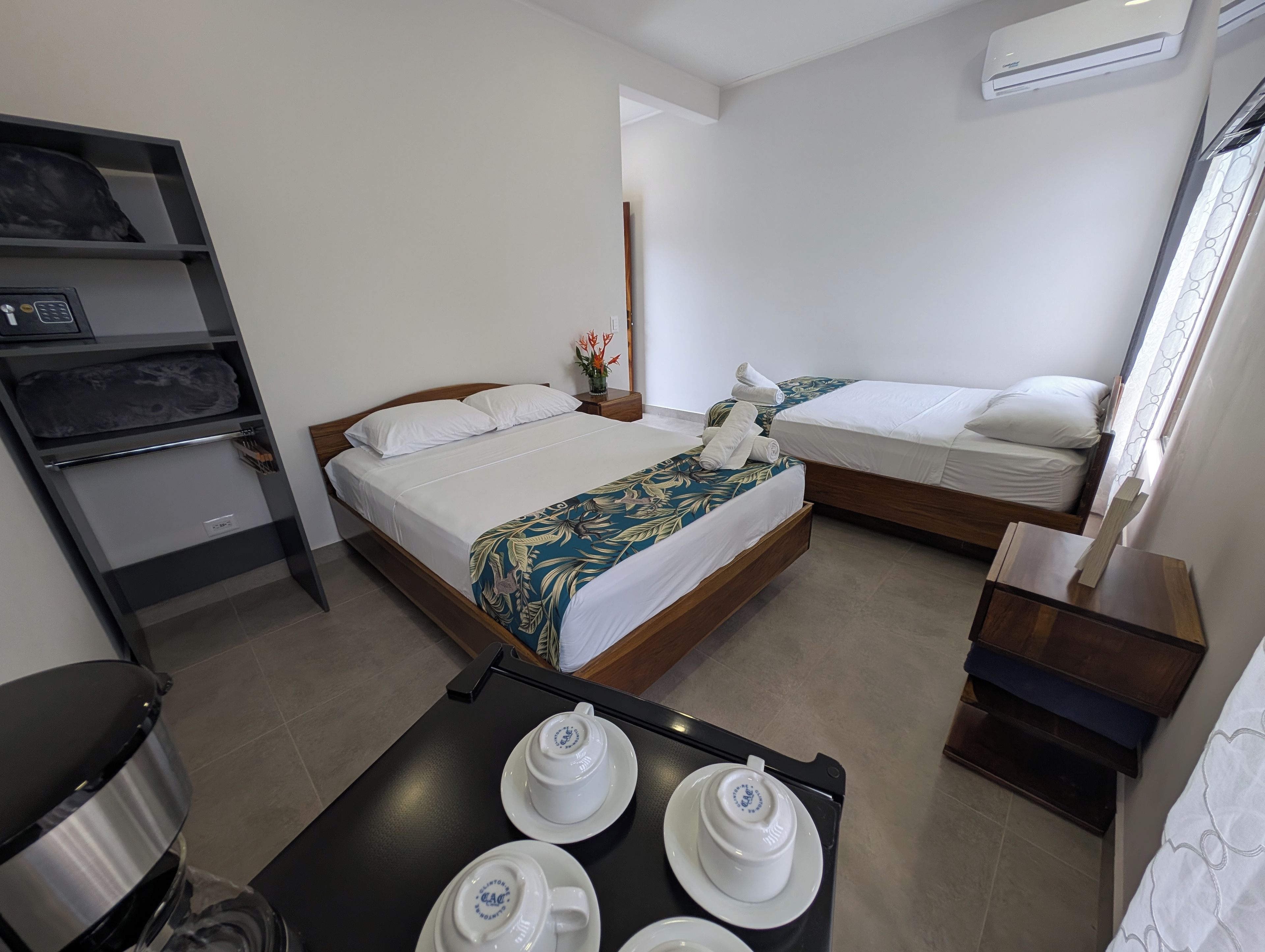 A hotel room with two beds, a small table with tea sets, a shelf with towels, and an air conditioning unit.