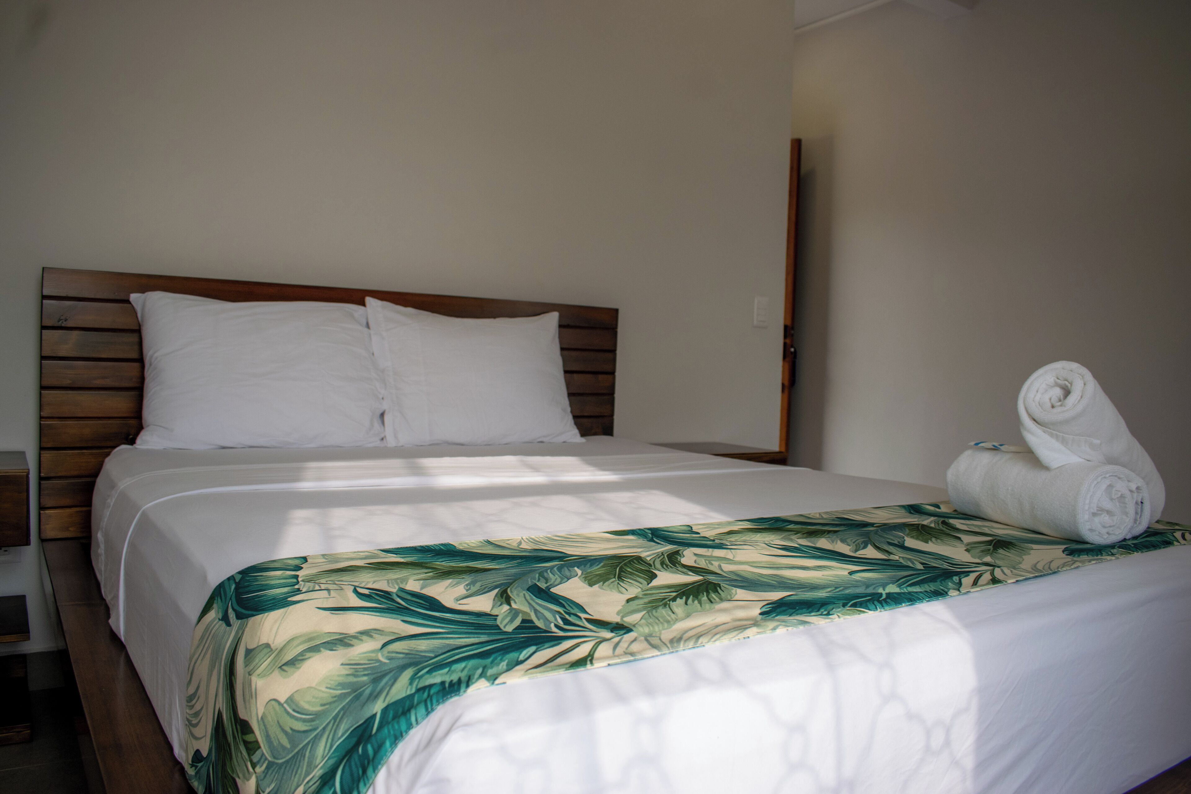 A neatly made bed with a wooden headboard, white bedding, and a tropical leaf-patterned throw.