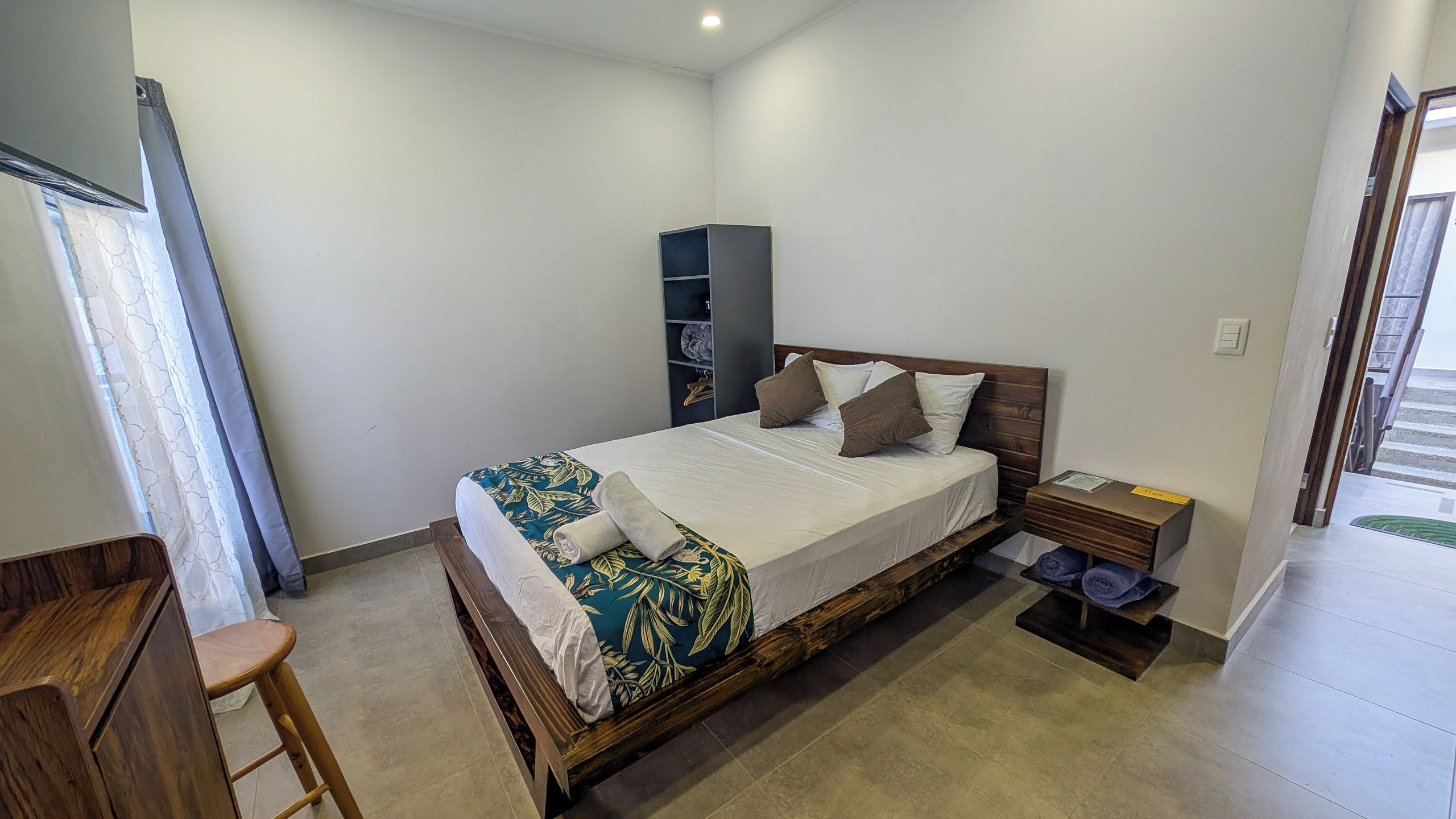 A bedroom with a bed, a nightstand, a wooden chair, a wardrobe, and a window with curtains.