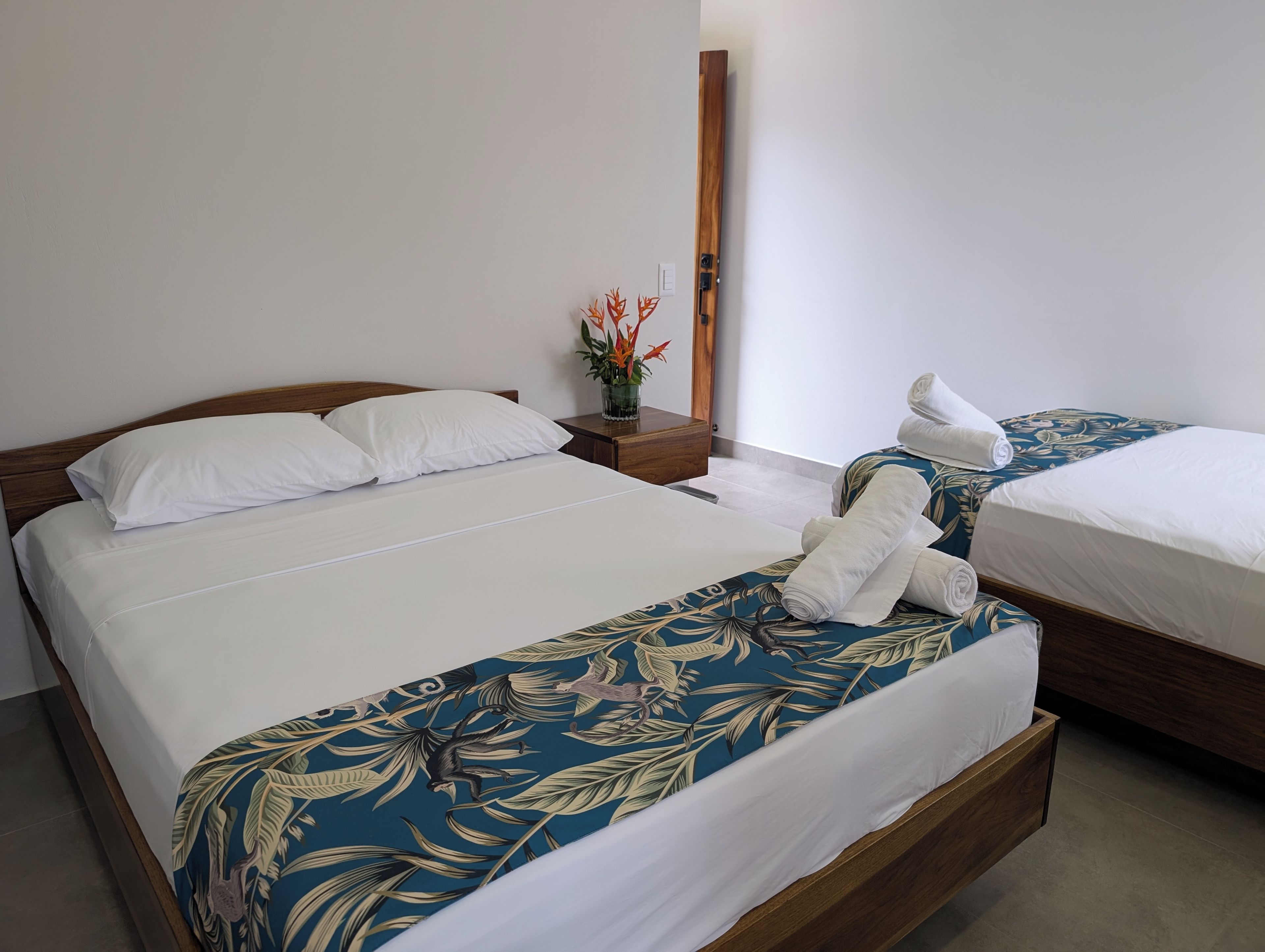 Una habitación de hotel con dos camas, cabecera de madera, mesita de noche con un florero y un espejo.