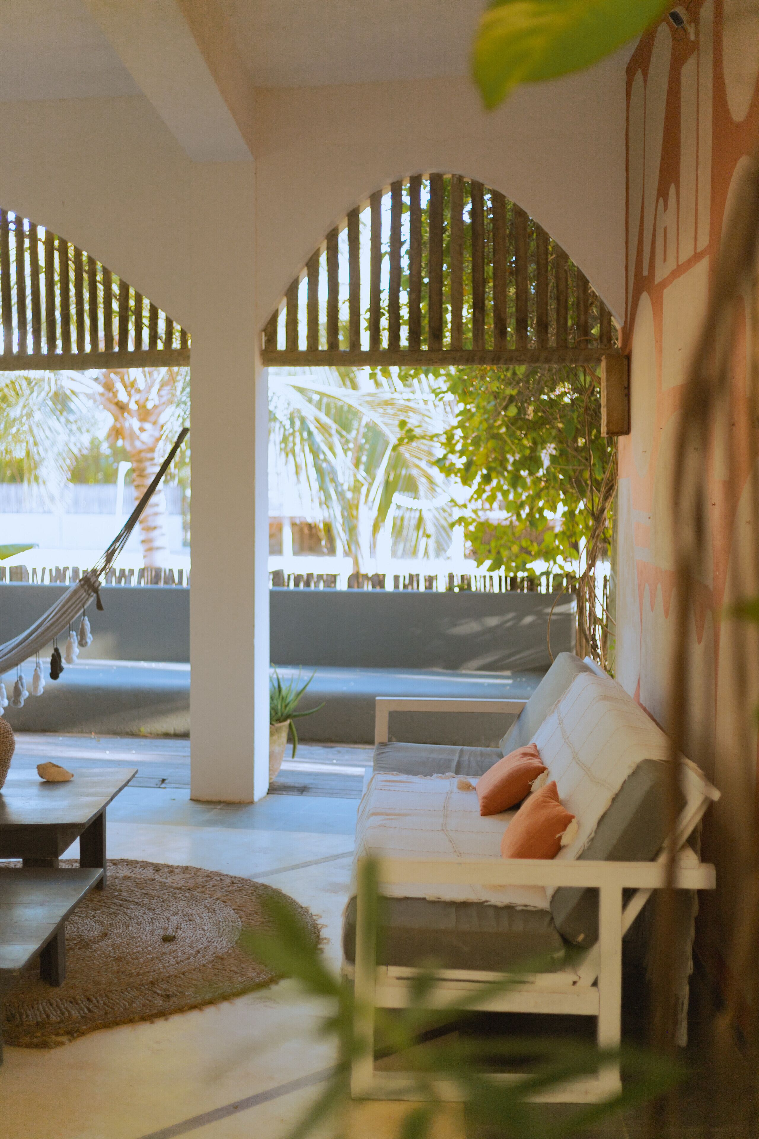 A hammock, a white sofa, and a small table in a covered outdoor area with a view of palm trees.