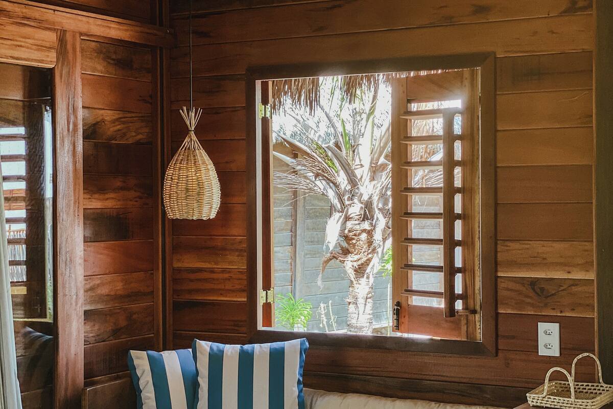 A wooden room with a bed, a woven light fixture, and a window with a view of palm trees.