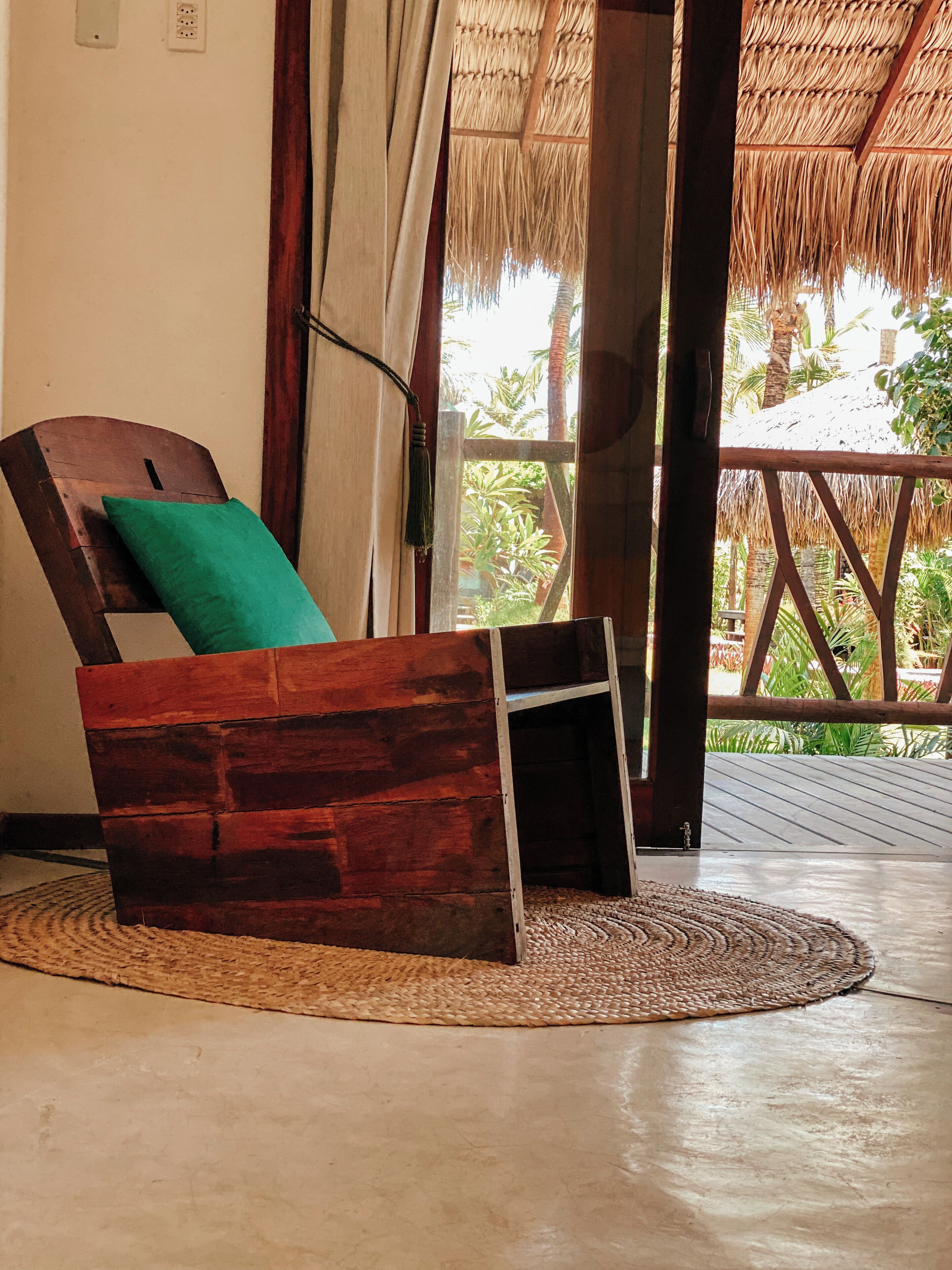 A wooden bench with a green pillow, a woven mat, and a view of a thatched-roof structure and greenery outside.