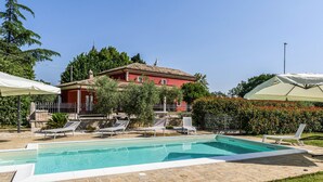 Une piscine entourée de chaises longues et de parasols, elle-même bordée d’un jardin et d’un bâtiment rouge à toit de tuiles.