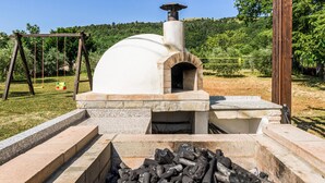 Un four à pizza au feu de bois, doté d’une base en pierre et d’une cheminée, entouré d’une zone herbeuse et d’arbres.