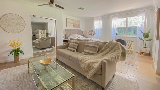 A modern living room with a glass coffee table, a beige sofa, a ceiling fan, and a large mirror. - Private Paradise - Large, Peaceful Studio w Patio (Naples)
