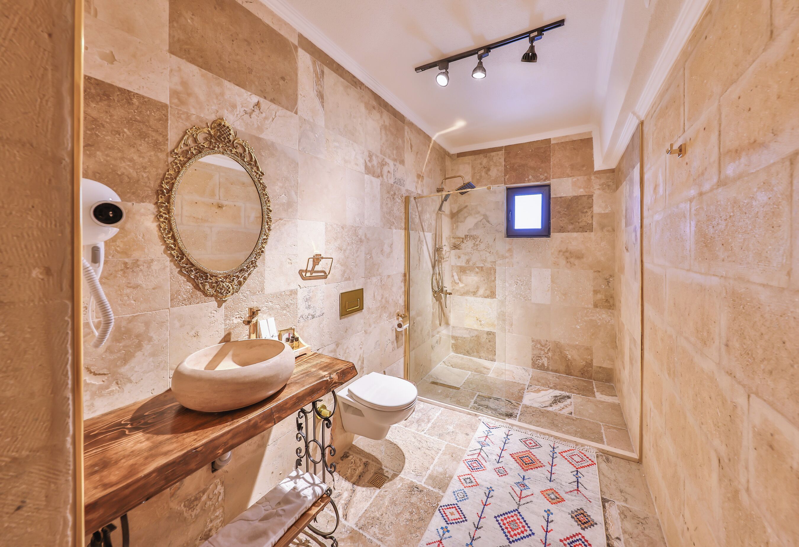 A bathroom with a marble wall, a wooden vanity, a white sink, a toilet, a shower with a glass door, and a patterned floor.