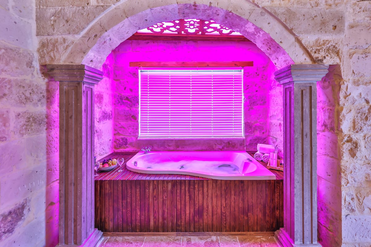Una habitación con paredes de piedra, una bañera de madera, una ventana con persianas y un techo decorativo.