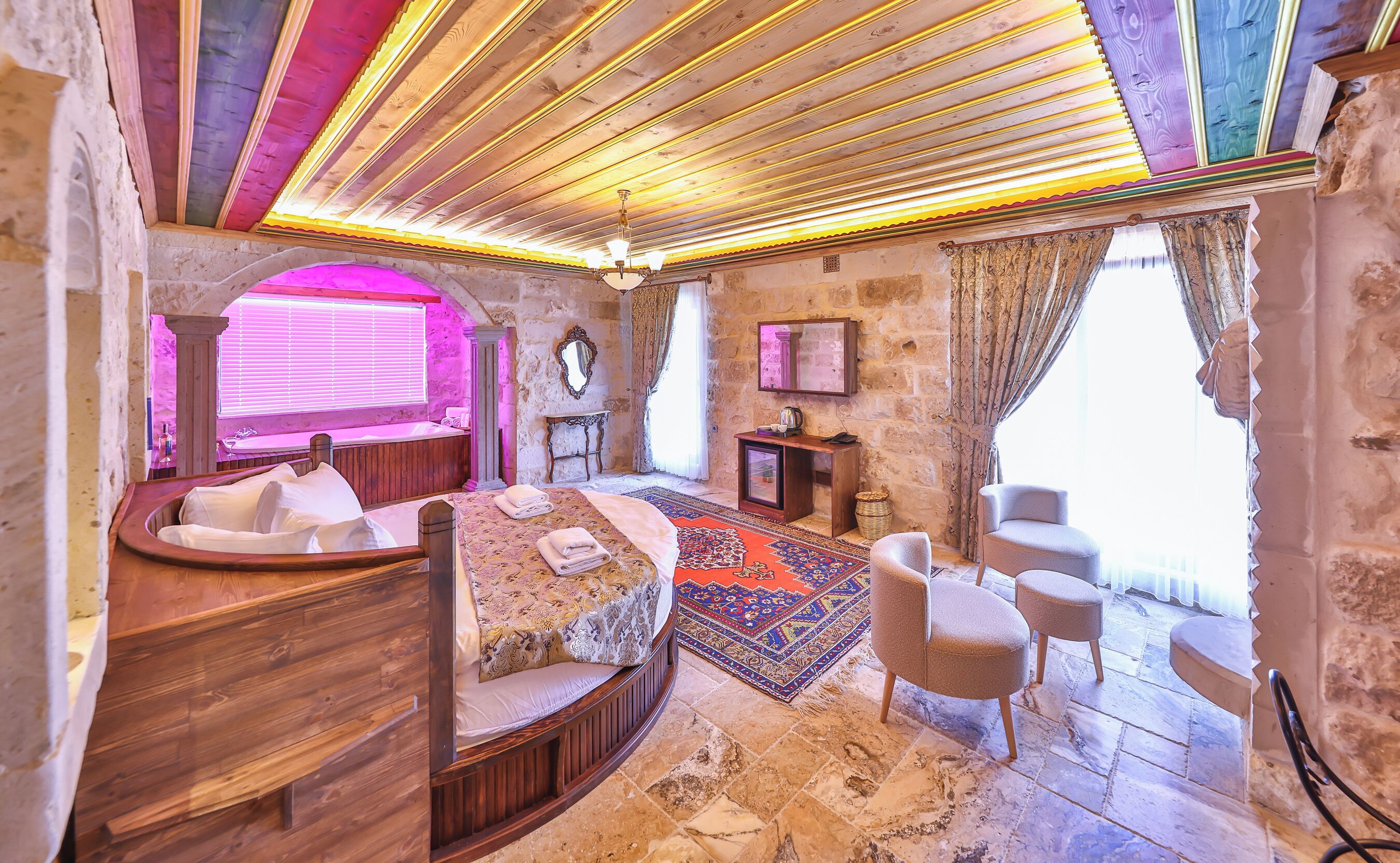 Une pièce avec un lit en bois, un petit canapé, une grande moquette, un mur en pierre et une fenêtre avec des rideaux.