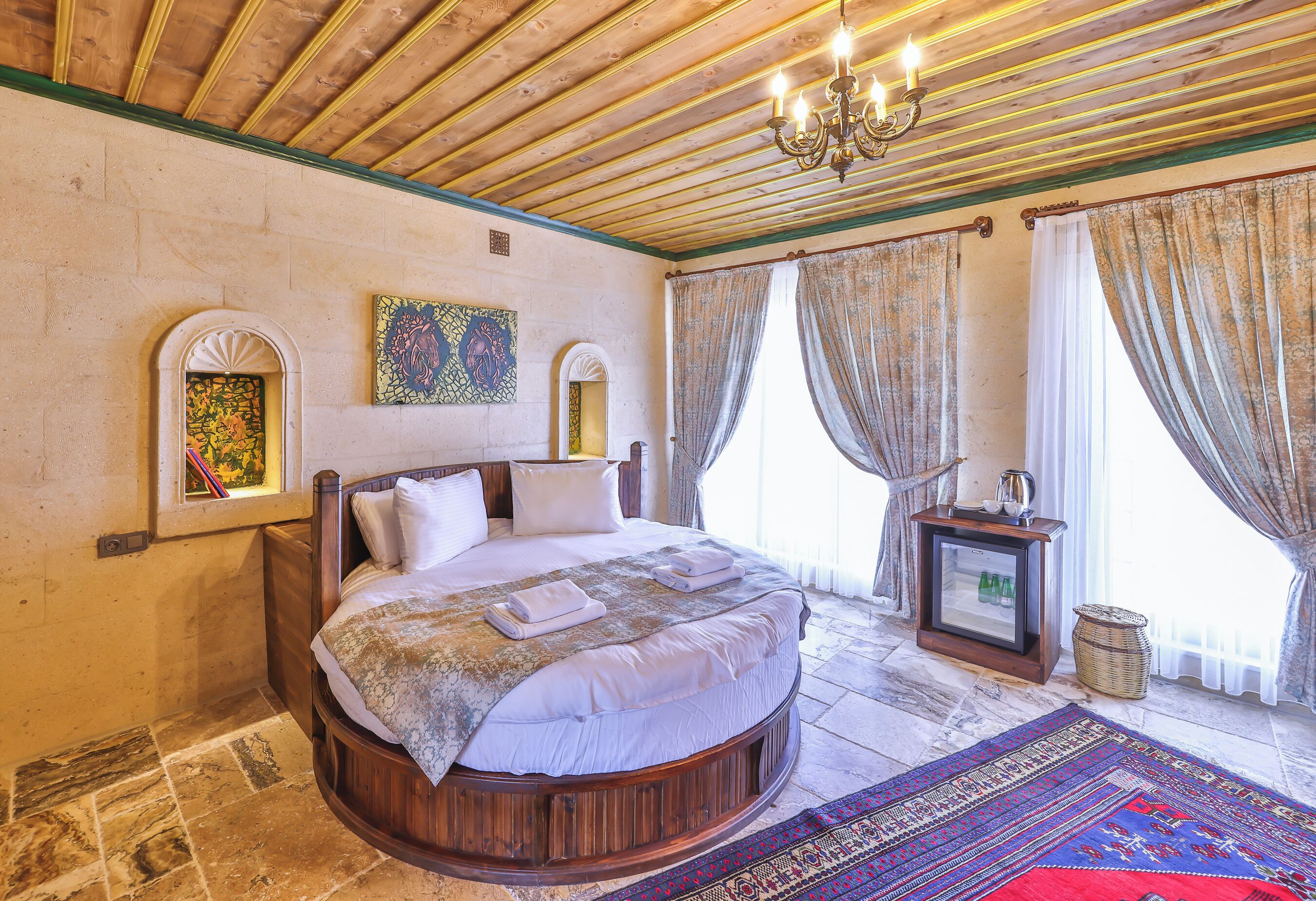 Ein Schlafzimmer mit einem runden Bett, einer Kristallleuchtre, einem kleinen Tisch mit Fernseher und einem Fenster mit Vorhängen.
