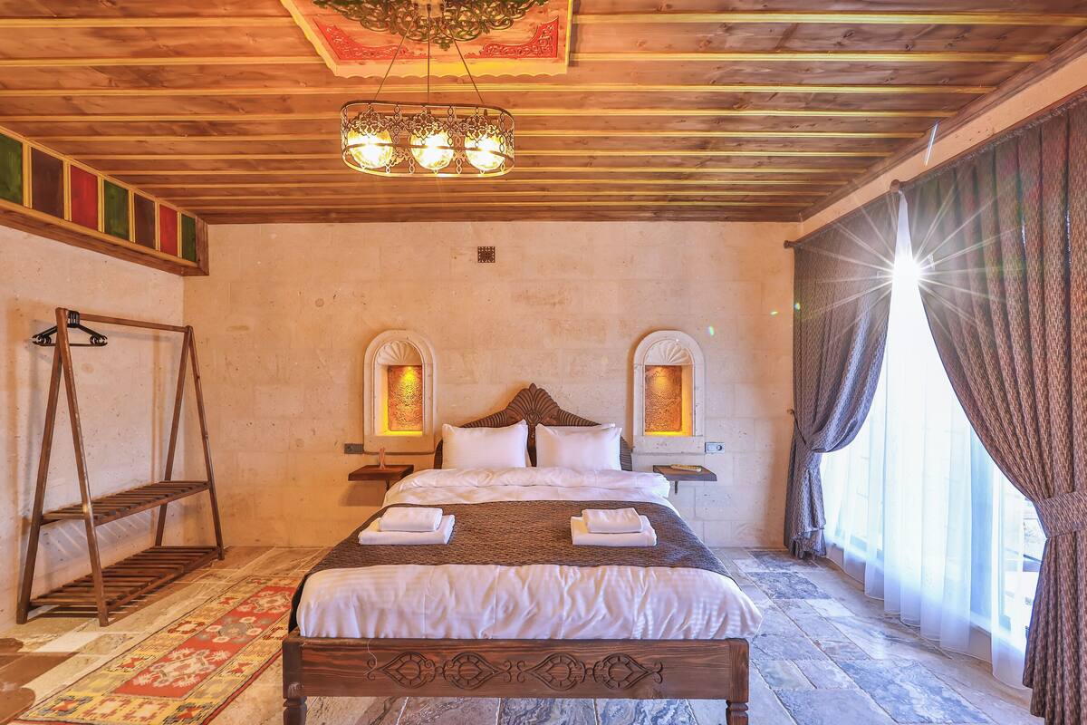 Una cama con estructura de madera, cabecera, dos mesitas de noche, lámpara colgante y una ventana con cortinas.