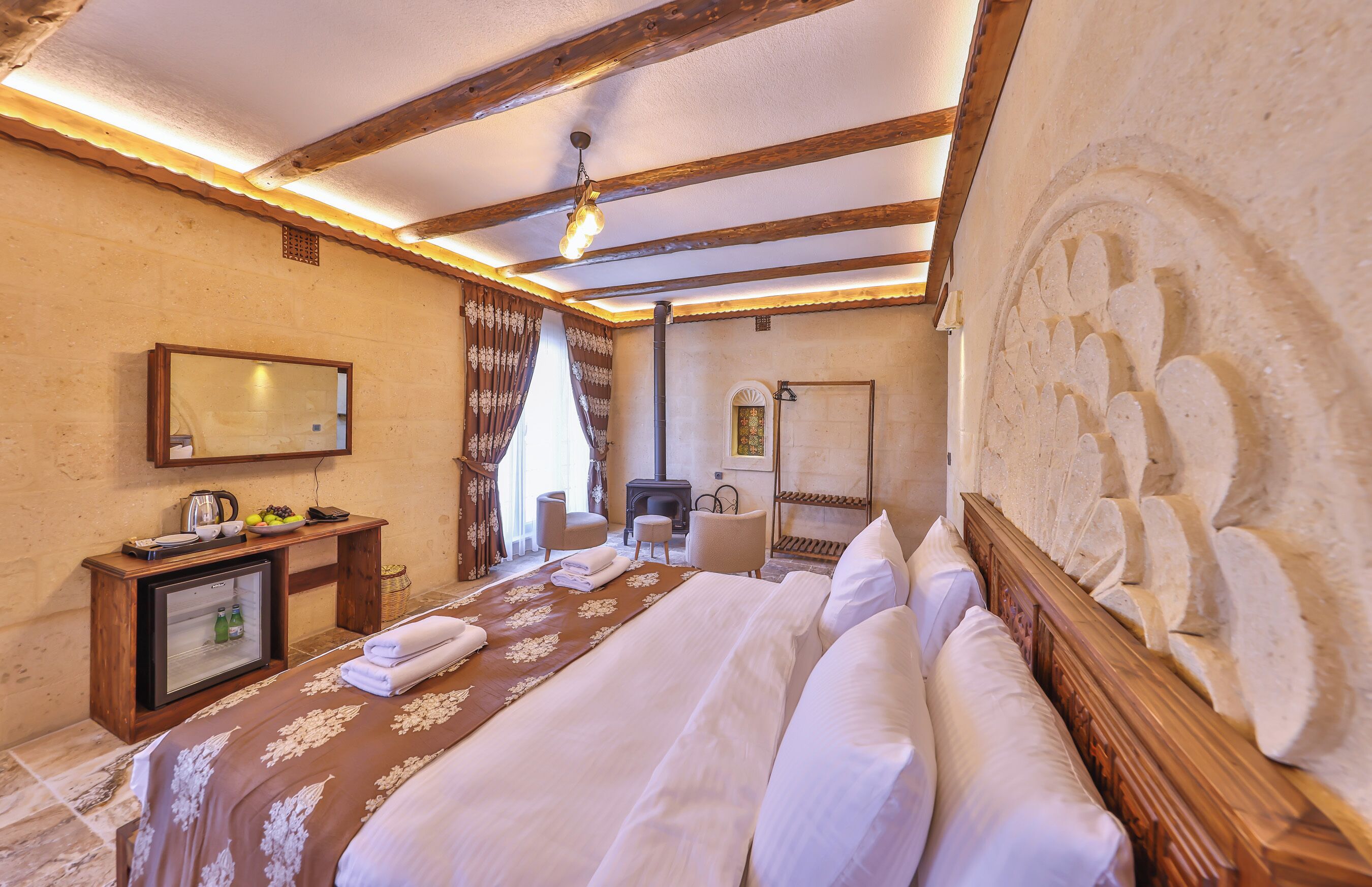A hotel room with a bed, a small desk with a refrigerator, a chair, a fireplace, and a large stone wall.