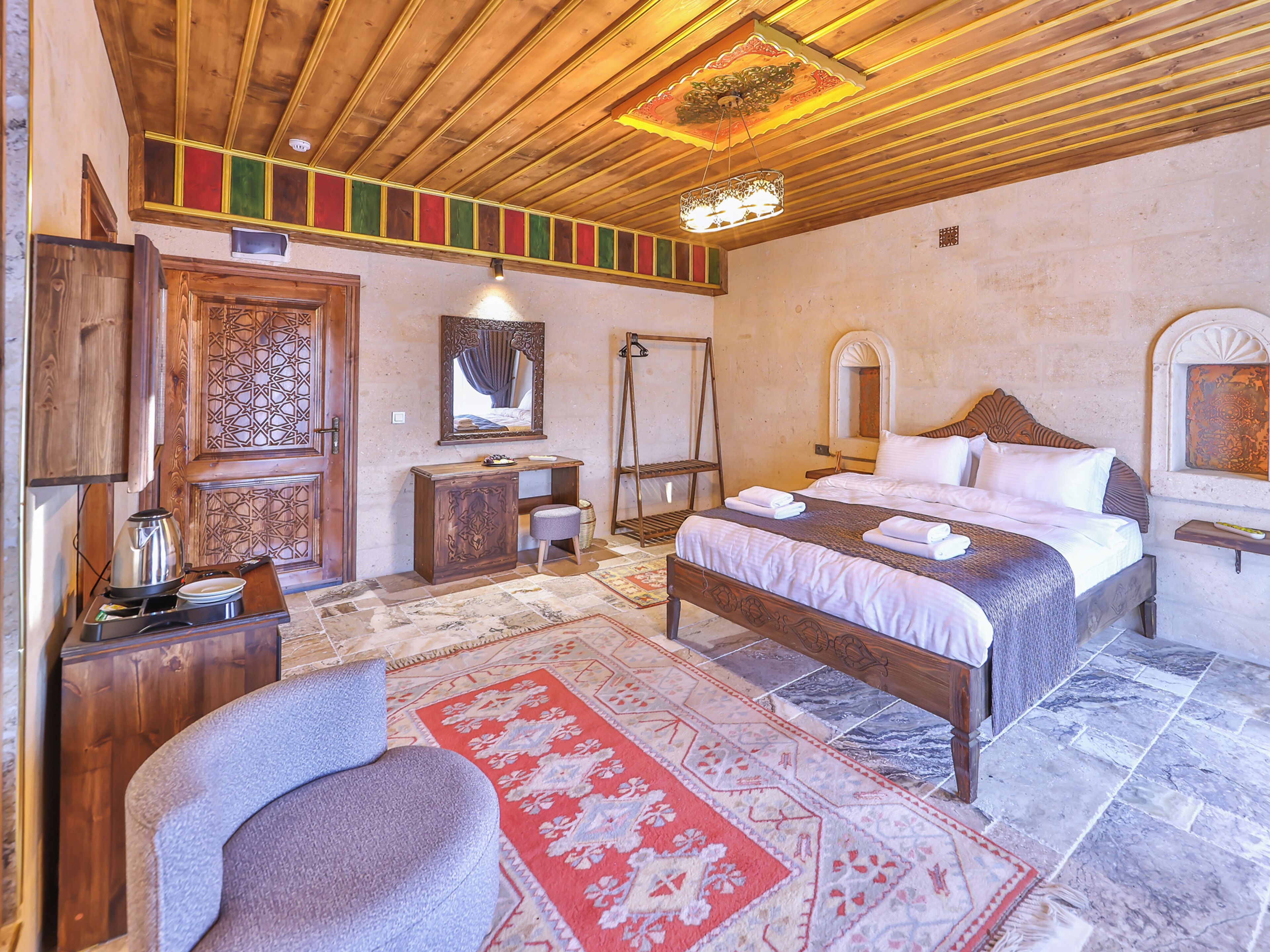 Un dormitorio con techo de madera, una cama con ropa de cama blanca, un aparador de madera, un espejo y una alfombra roja con estampado.