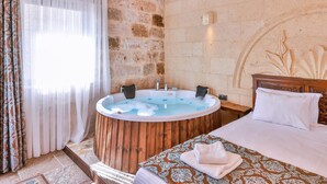 A room with a wooden bathtub, a bed with white linens, a stone wall, and a window with sheer curtains.