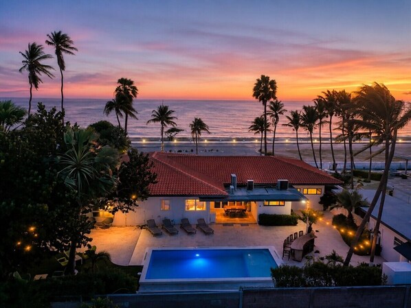 A beachfront property with a swimming pool, lounge chairs, and a red-roofed building.
