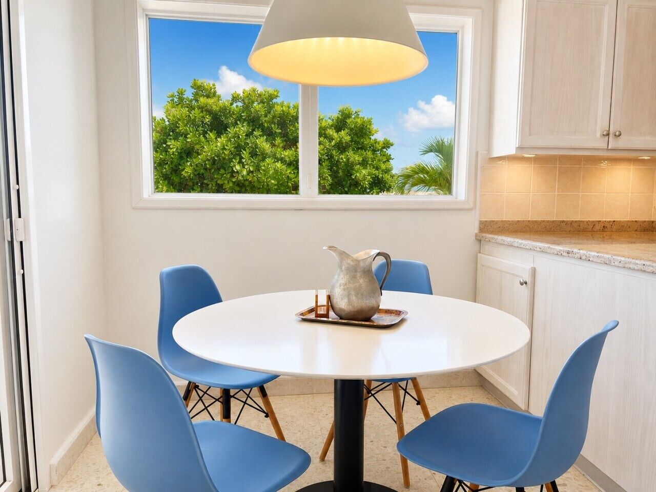 A round table with four chairs, a pitcher, and a window with a view of trees.