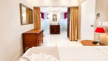 A hotel room with a bed, a wooden dresser, a vanity with a mirror, a red lamp, and a view of the bathroom.