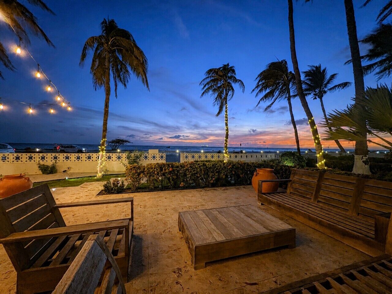 An outdoor seating area with wooden furniture and palm trees, overlooking a body of water.