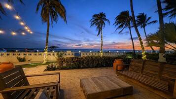 An outdoor seating area with wooden furniture and palm trees, overlooking a body of water.