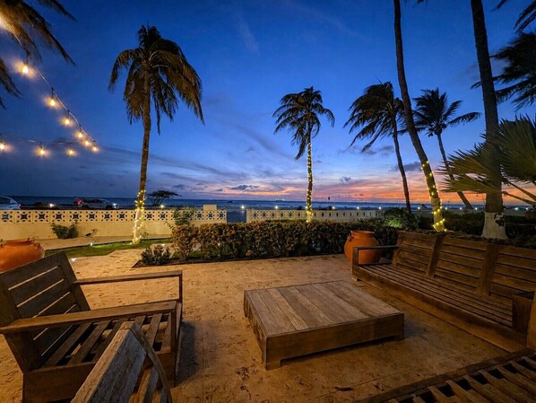 Un espacio al aire libre con mobiliario de madera y palmeras, con vista a una masa de agua.