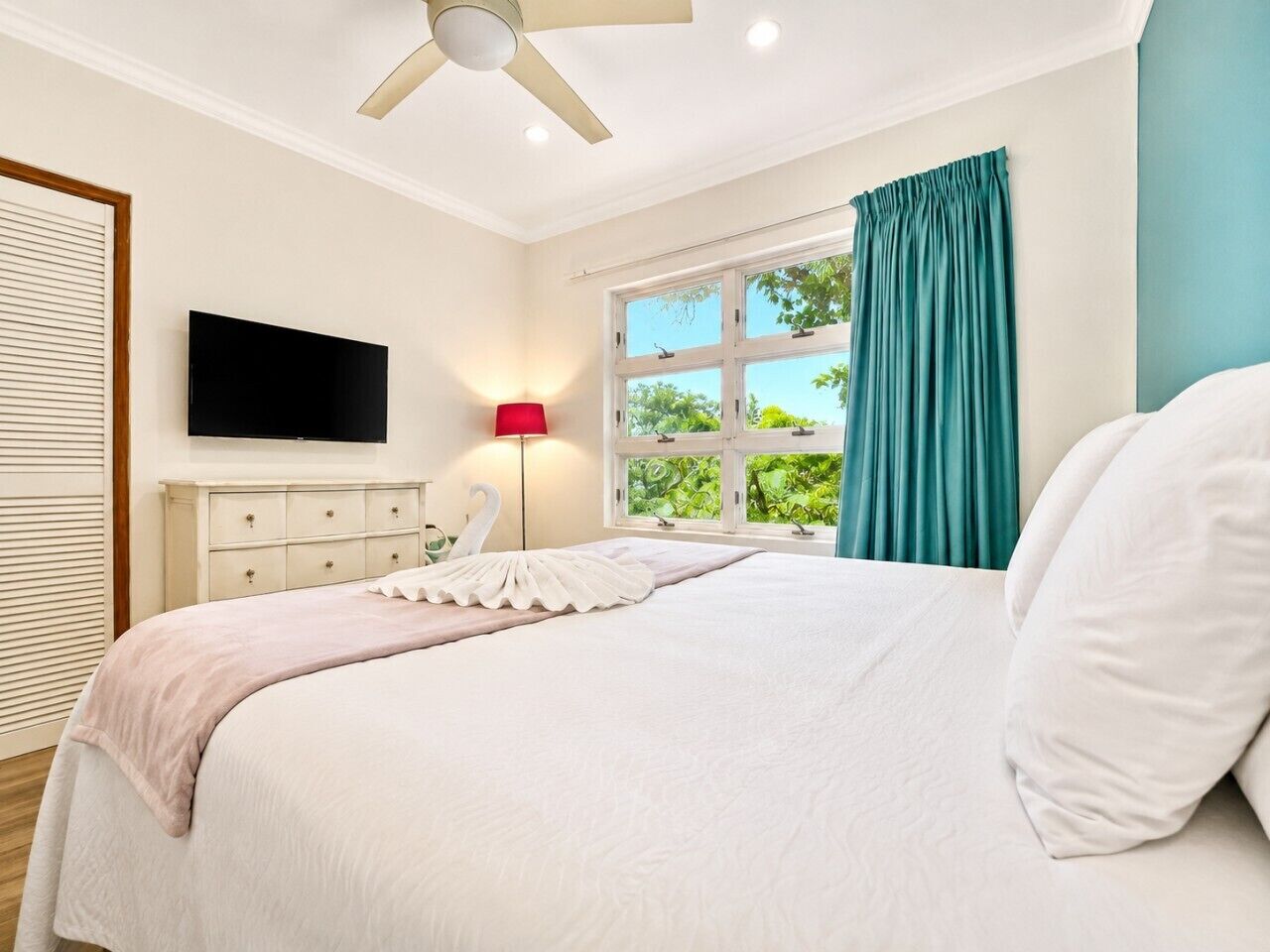 A bedroom with a bed, a flat-screen TV, a bedside lamp, a window with curtains, and a ceiling fan.
