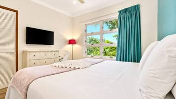 A bedroom with a bed, a flat-screen TV, a bedside lamp, a window with curtains, and a ceiling fan.