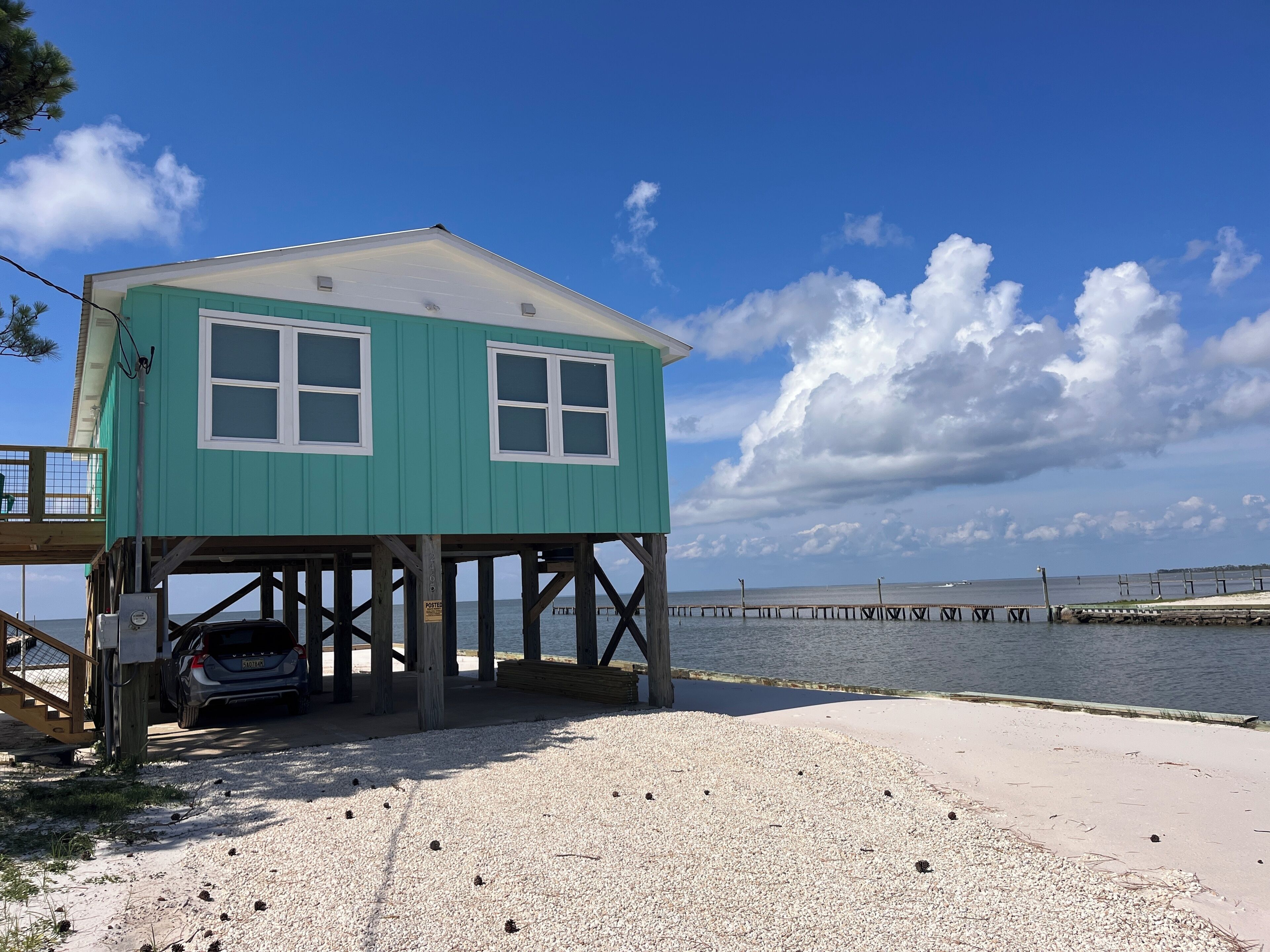 Un bâtiment turquoise avec une voiture garée en dessous, situé sur une plage de galets, avec une jetée qui s’avance dans l’eau.