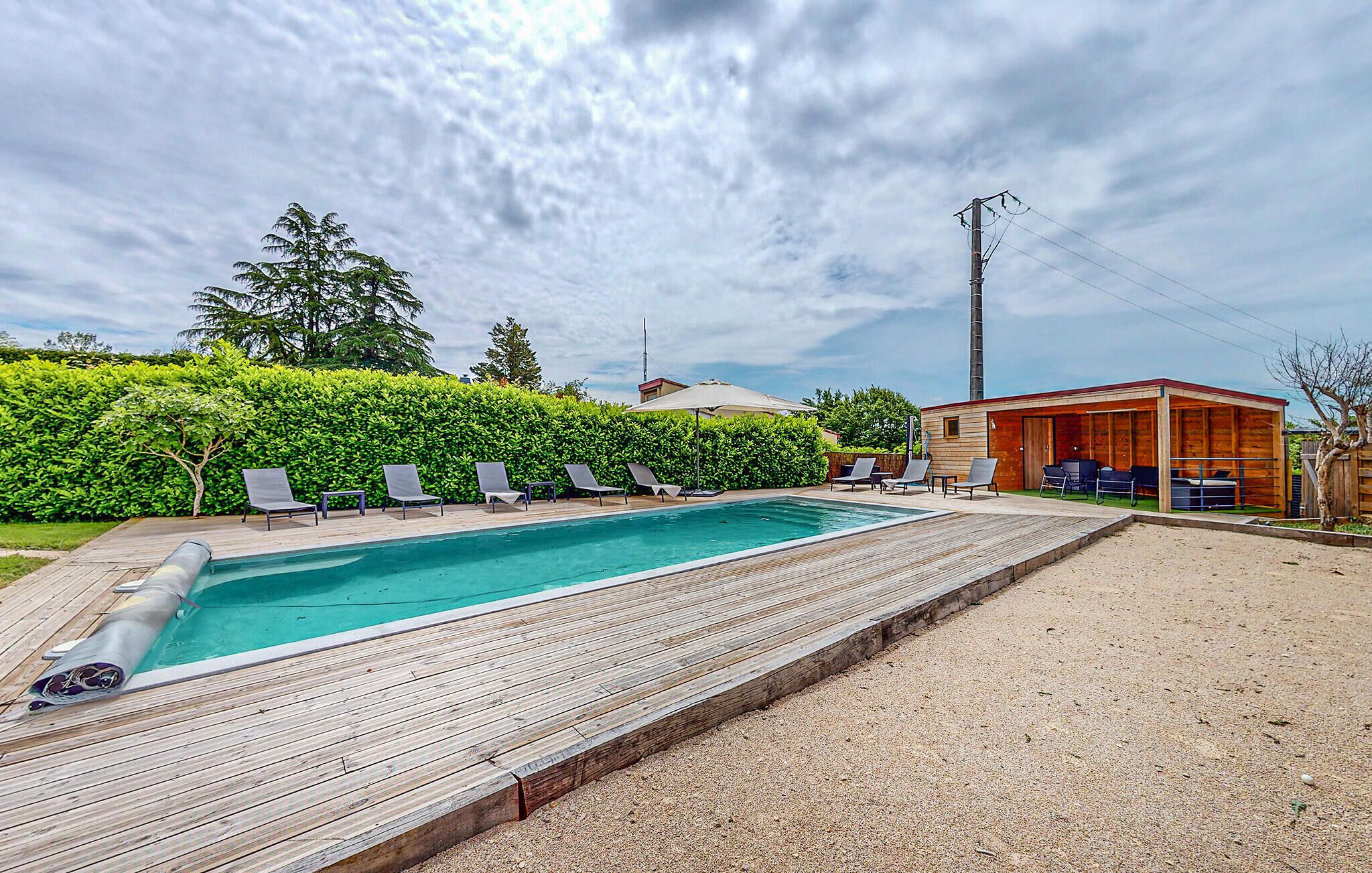 Une piscine rectangulaire dotée d’une terrasse en bois, entourée d’une haie et bordée d’un bâtiment en bois avec des sièges extérieurs.