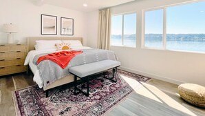 Une chambre à coucher comprenant un lit, une table de chevet, un banc, de grandes fenêtres et une vue sur une étendue d’eau.