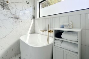 A modern bathroom with a marble wall, a freestanding bathtub, and a white vanity with shelves.