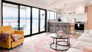 Un salon moderne avec un canapé jaune, une table basse ronde et une grande porte-fenêtre coulissante donnant sur un balcon avec vue sur l’eau.