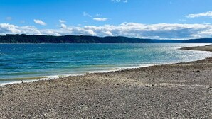 Une plage de galets bordée d’une eau calme, avec des collines boisées à l’horizon et un ciel dégagé.