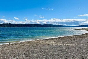 A pebbly beach with calm water, distant forested hills, and a clear sky.