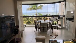 A balcony with a view of the sea, a dining area with chairs, a kitchen with a counter, and a TV on a stand. - Riviera: M3 Kingston Town 61, beach front (Bertioga)
