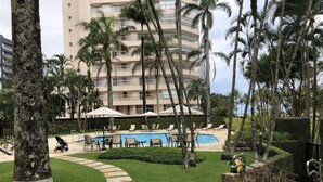 A pool area with lounge chairs, a pathway, and a building in the background. - Riviera: M3 Kingston Town 61, beach front (Bertioga)