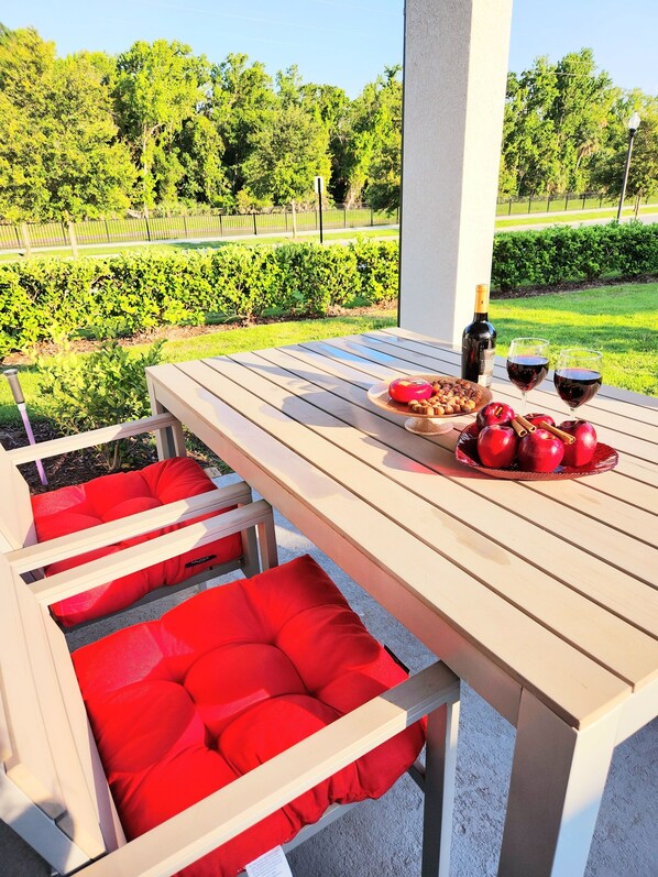 An outdoor dining area with a wooden table, red cushioned chairs, a bottle of wine, glasses, and a plate of food. - Brand New Modern, Luxury Villa with Resort Pools near Disney! (Kissimmee)